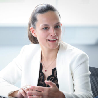 Eine Frau in einem weißen Blazer sitzt an einem Tisch, spricht und gestikuliert mit der Hand; vor ihr steht eine Kaffeetasse.