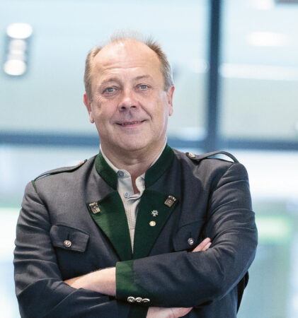 Ein Mann in Blazer, grünem Hemd, khakifarbenen Shorts und Wanderschuhen steht selbstbewusst in der Lobby eines modernen, hell erleuchteten Büros.