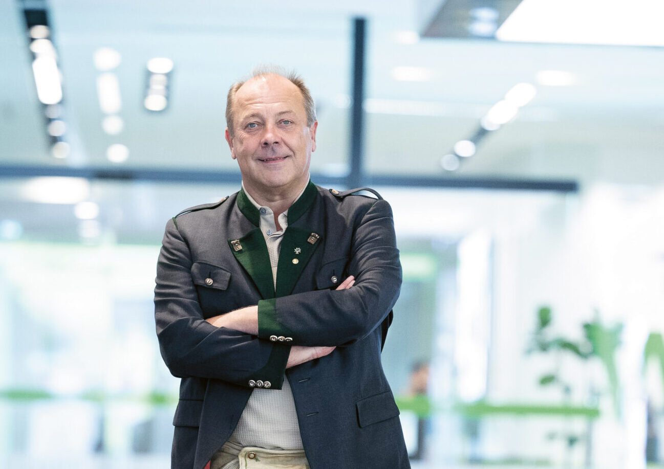 Ein Mann in Blazer, grünem Hemd, khakifarbenen Shorts und Wanderschuhen steht selbstbewusst in der Lobby eines modernen, hell erleuchteten Büros.