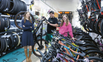 Drei Personen stehen lächelnd in einem Fahrradgeschäft, umgeben von Fahrrädern und Fahrradzubehör, das auf Gestellen und Regalen ausgestellt ist.