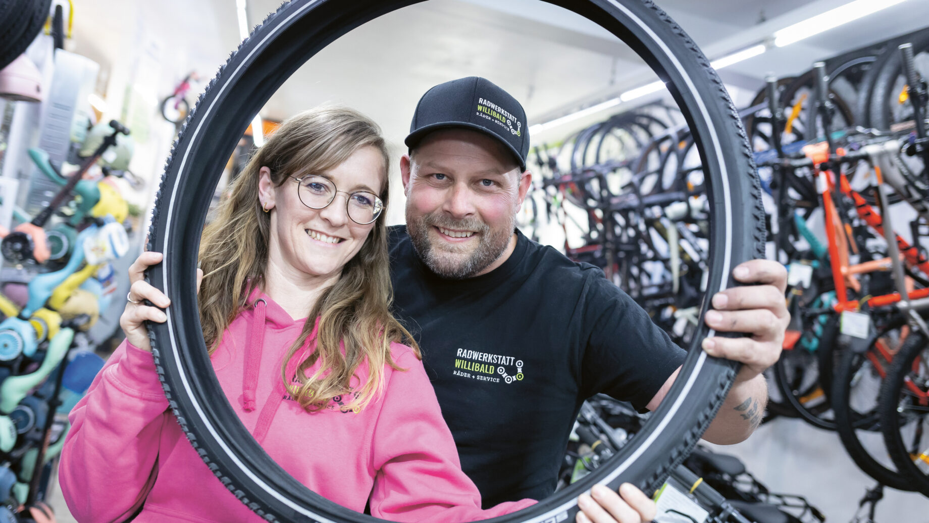 Zwei Menschen, die lächelnd gemeinsam posieren, während sie einen Fahrradreifen hochhalten – in einem Fahrradgeschäft voller Fahrräder und Fahrradzubehör.