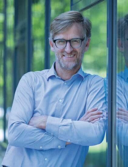 Ein lächelnder Mann mit Brille und Bart, der ein hellblaues Hemd trägt, steht mit verschränkten Armen da; in der Glaswand neben ihm ist sein Spiegelbild zu sehen.