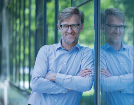 Ein lächelnder Mann mit Brille und Bart, der ein hellblaues Hemd trägt, steht mit verschränkten Armen da; in der Glaswand neben ihm ist sein Spiegelbild zu sehen.