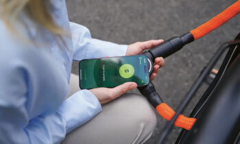 Eine Person hält ein Smartphone mit entsperrtem Bildschirm in die Nähe eines orangefarbenen Fahrradschlosses und tut so, als ob sie das Telefon zum Aufschließen des Fahrrads verwenden würde.