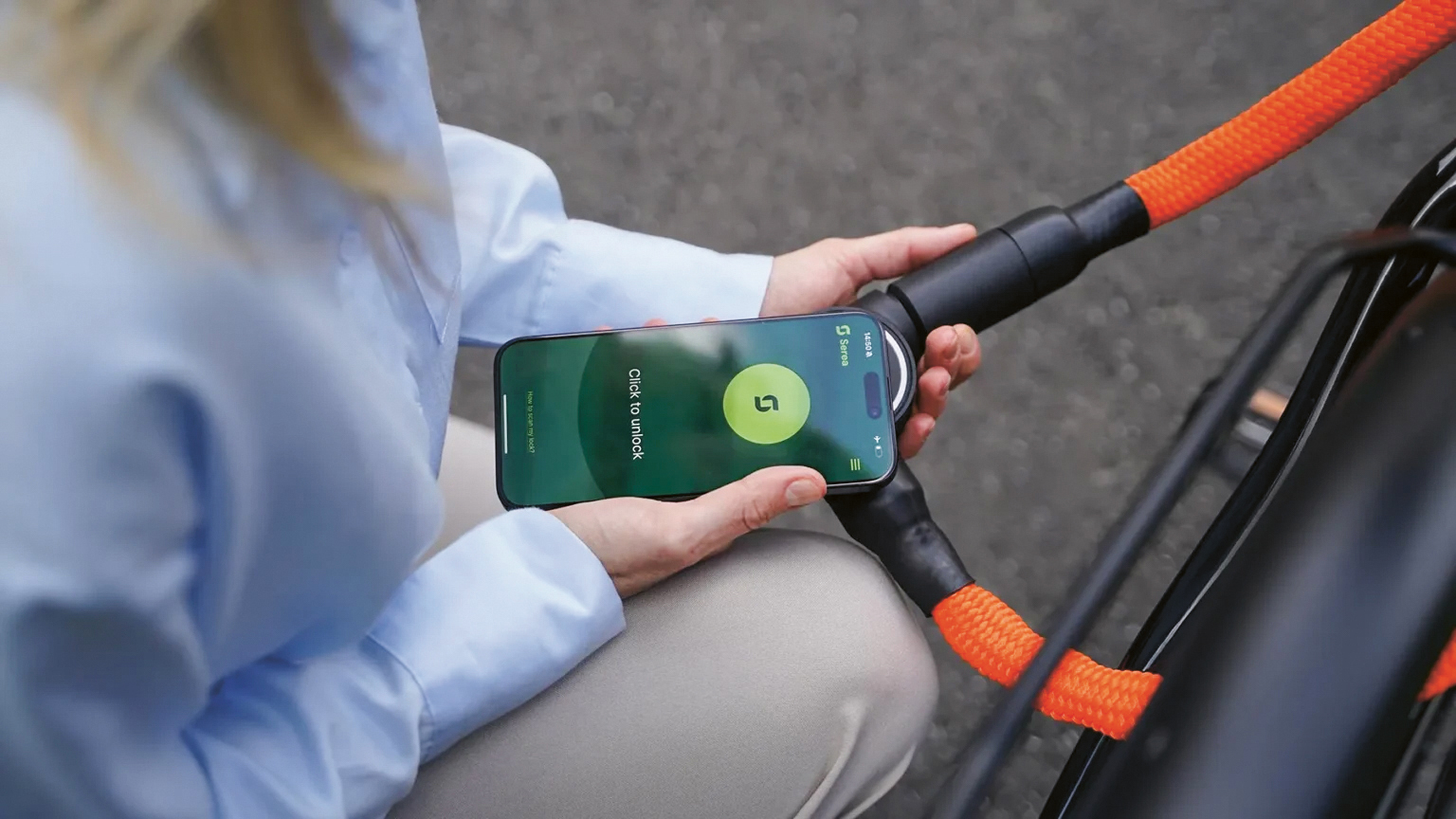 Eine Person hält ein Smartphone mit entsperrtem Bildschirm in die Nähe eines orangefarbenen Fahrradschlosses und tut so, als ob sie das Telefon zum Aufschließen des Fahrrads verwenden würde.