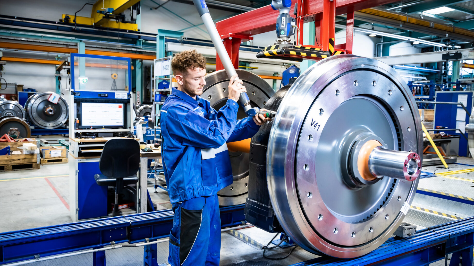 Ein Fabrikarbeiter in blauer Arbeitskleidung montiert ein großes Metallrad und feiert damit an der Fertigungsstraße „125 Jahre Siemens in der Steiermark“.