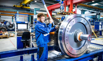 Ein Fabrikarbeiter in blauer Arbeitskleidung montiert ein großes Metallrad und feiert damit an der Fertigungsstraße „125 Jahre Siemens in der Steiermark“.