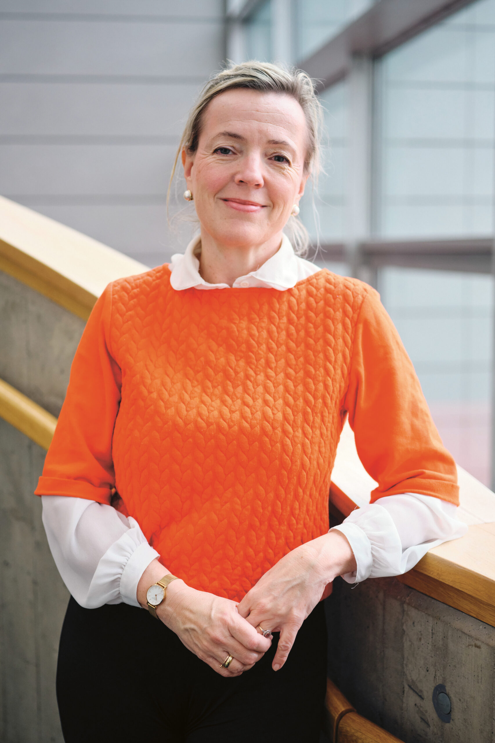 Eine Frau in einem orangefarbenen, strukturierten Oberteil und einer weißen Bluse lächelt, während sie in einem Innenraum neben einer Treppe steht, im Hintergrund sind große Fenster zu sehen.