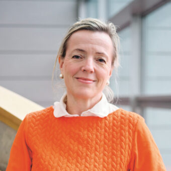 Eine Frau mit hellem Haar, die ein orangefarbenes, strukturiertes Oberteil und eine weiße Bluse trägt, steht lächelnd in einem Raum und stützt sich mit der Hand auf das Treppengeländer.