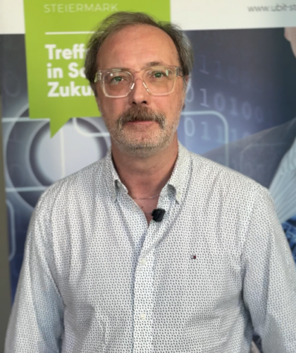 Ein Mann mit Brille und grauem Bart steht neben einem Schild von UBIT Steiermark und ist bereit, seine Erkenntnisse zu teilen.