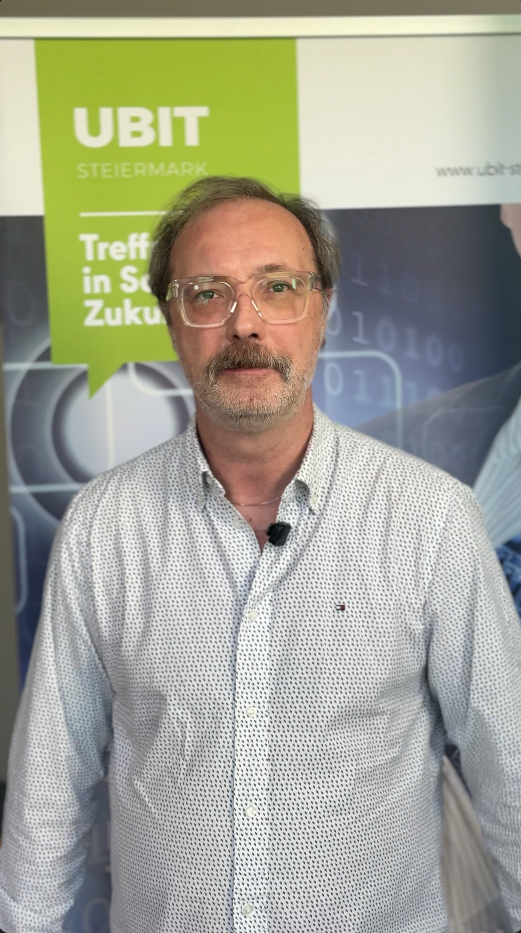 Ein Mann mit Brille und grauem Bart steht neben einem Schild von UBIT Steiermark und ist bereit, seine Erkenntnisse zu teilen.