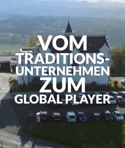 Ein großes Gebäude, vor dem viele Autos geparkt sind, inmitten einer malerischen Landschaft, überlagert von dem Text „Vom Traditionsbetrieb zum Global Player"
