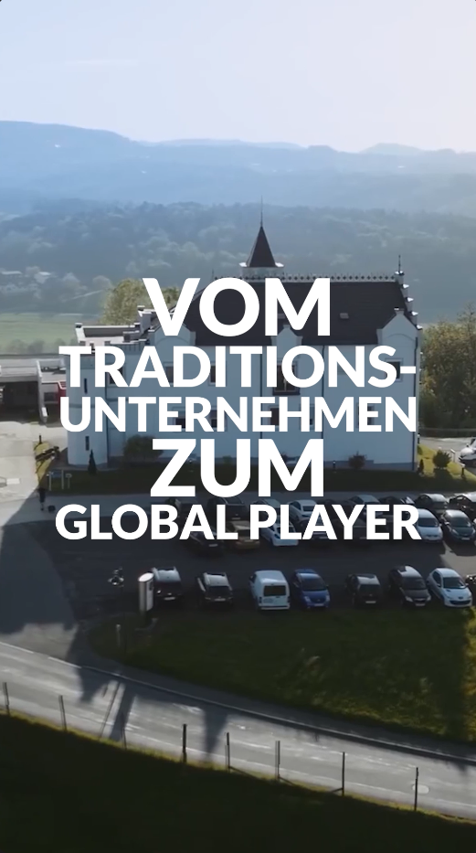 Ein großes Gebäude, vor dem viele Autos geparkt sind, inmitten einer malerischen Landschaft, überlagert von dem Text „Vom Traditionsbetrieb zum Global Player"