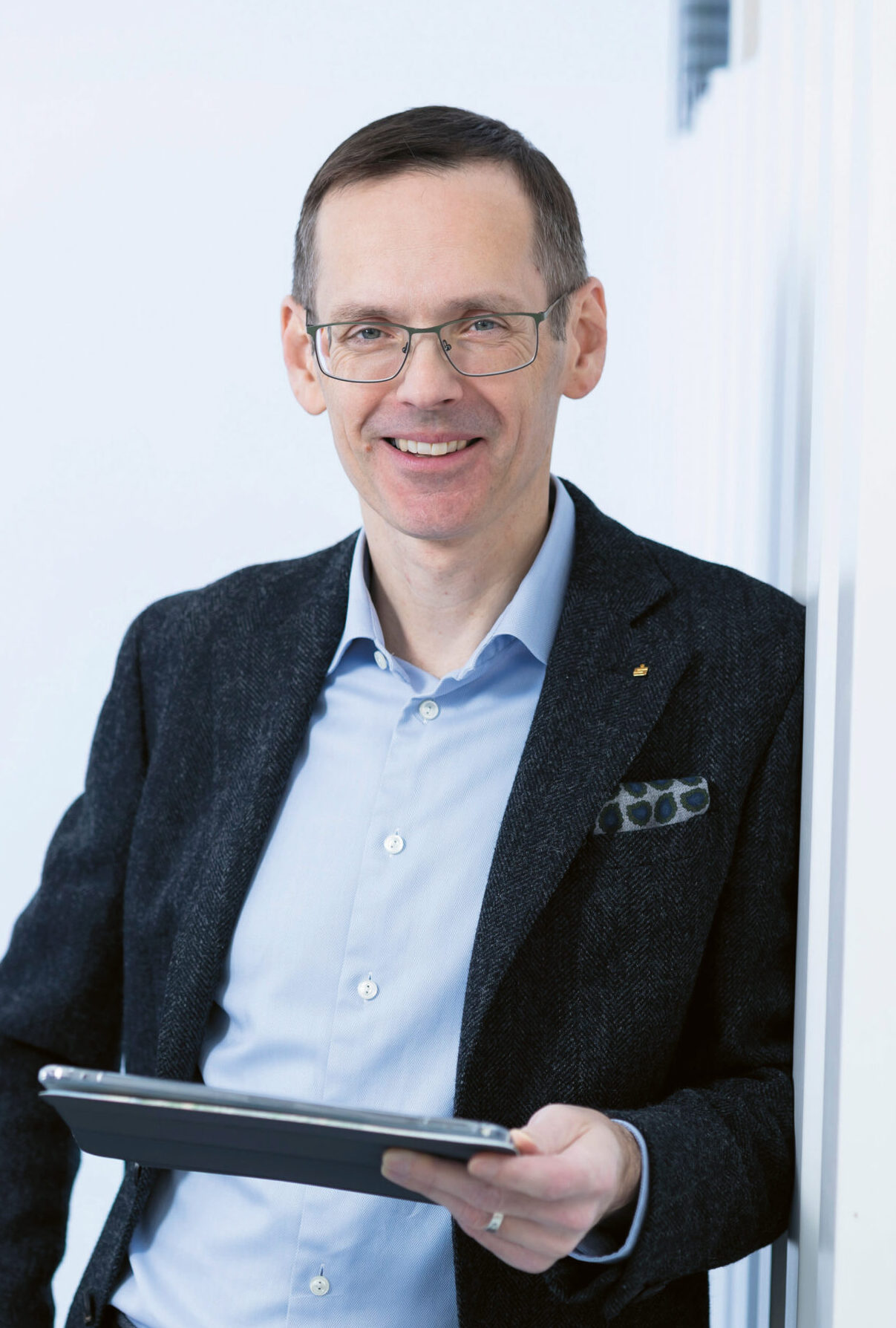 Ein lächelnder Mann mit Brille, der einen Blazer und ein hellblaues Hemd trägt, steht vor einer weißen Wand in einem hellen Büro und hält ein Tablet in der Hand.