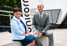 Eine Frau und ein Mann in Geschäftskleidung lächeln im Freien vor einem Banner mit der Aufschrift „Unicorn Science Meets Business“ und unterhalten sich über den Vergabeleitfaden der WKO Steiermark.