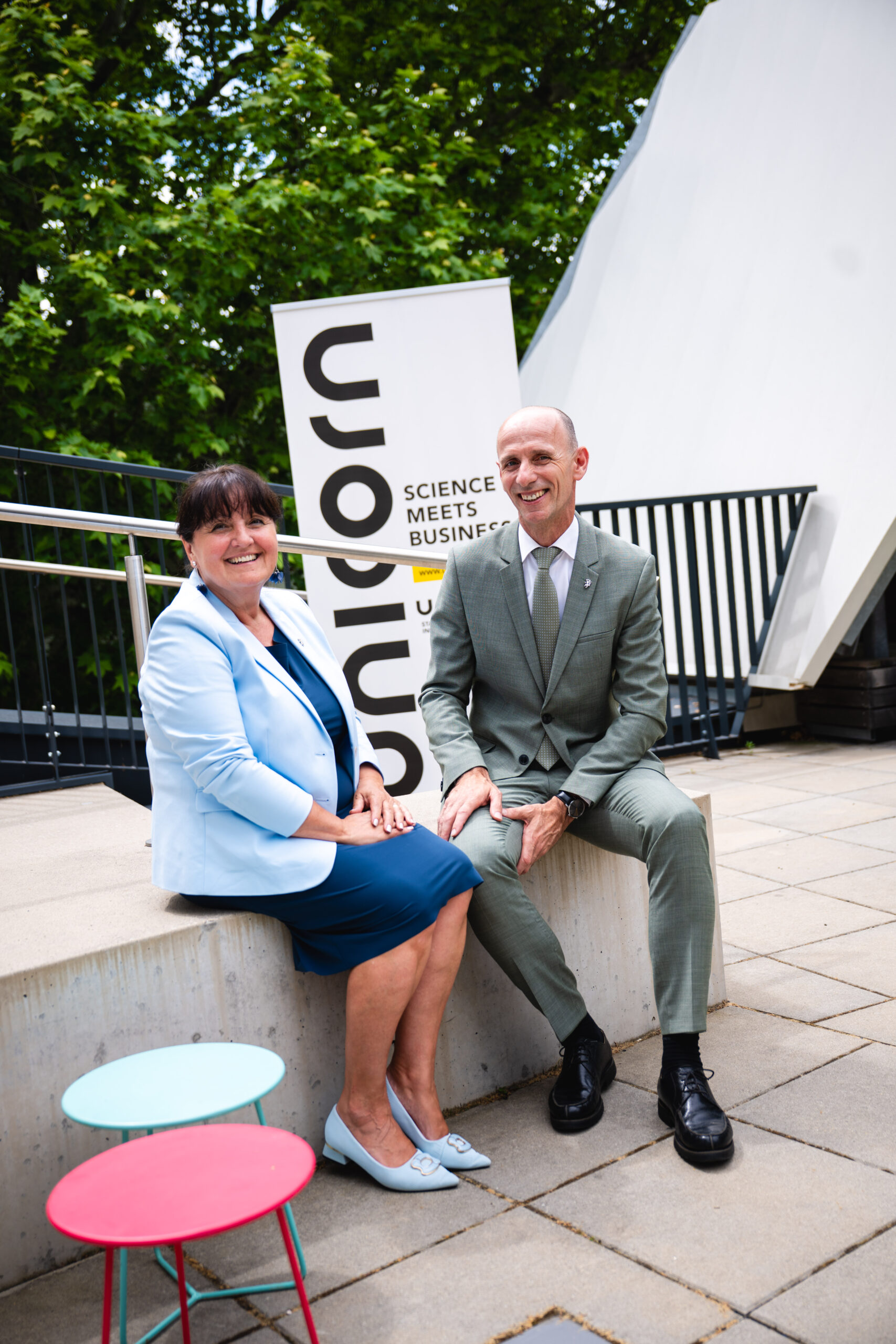 Eine Frau und ein Mann in Geschäftskleidung lächeln im Freien vor einem Banner mit der Aufschrift „Unicorn Science Meets Business“ und unterhalten sich über den Vergabeleitfaden der WKO Steiermark.