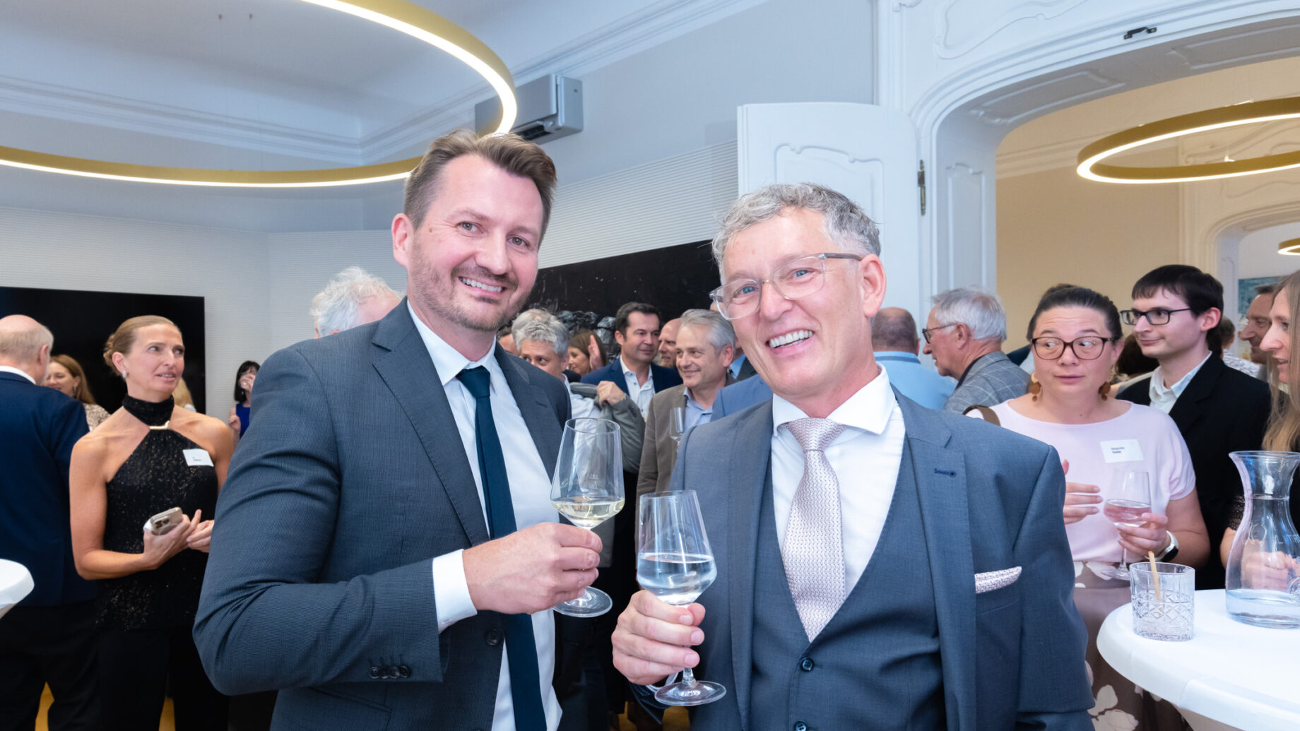 Zwei Männer in Anzügen lächeln und stoßen mit Weingläsern bei einer formellen Veranstaltung an, umgeben von anderen Personen, die in einem hellen, eleganten Raum verkehren.