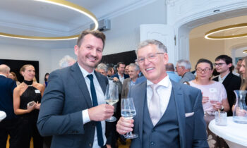 Zwei Männer in Anzügen lächeln und stoßen mit Weingläsern bei einer formellen Veranstaltung an, umgeben von anderen Personen, die in einem hellen, eleganten Raum verkehren.