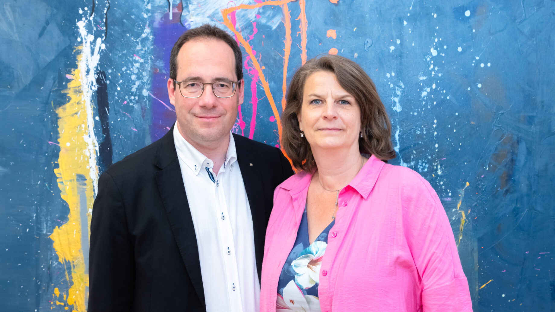 Ein Mann in einer schwarzen Jacke und eine Frau in einem rosa Hemd stehen zusammen vor einem bunten abstrakten blauen Hintergrund.