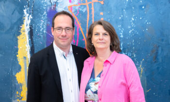 Ein Mann in einer schwarzen Jacke und eine Frau in einem rosa Hemd stehen zusammen vor einem bunten abstrakten blauen Hintergrund.