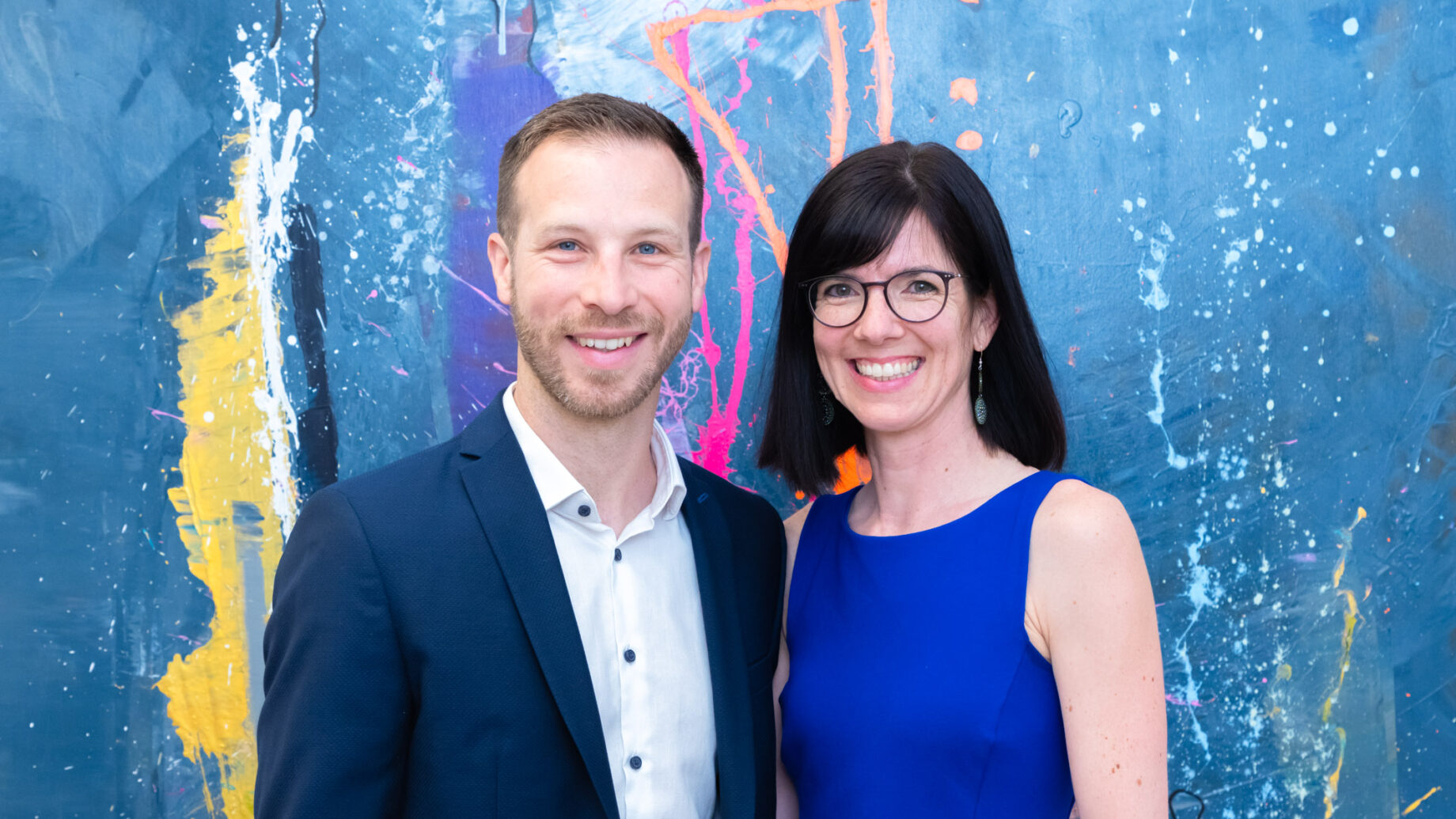 Ein Mann im Anzug und eine Frau in einem blauen Kleid lächeln vor einem bunten, abstrakt gemalten Hintergrund.