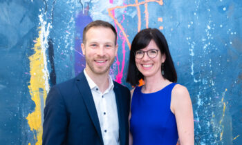 Ein Mann im Anzug und eine Frau in einem blauen Kleid lächeln vor einem bunten, abstrakt gemalten Hintergrund.
