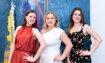 Drei Frauen lächeln und posieren vor einem farbenfrohen abstrakten Gemälde; eine trägt rot, eine andere weiß, und die dritte trägt ein schwarzes Blumenkleid.