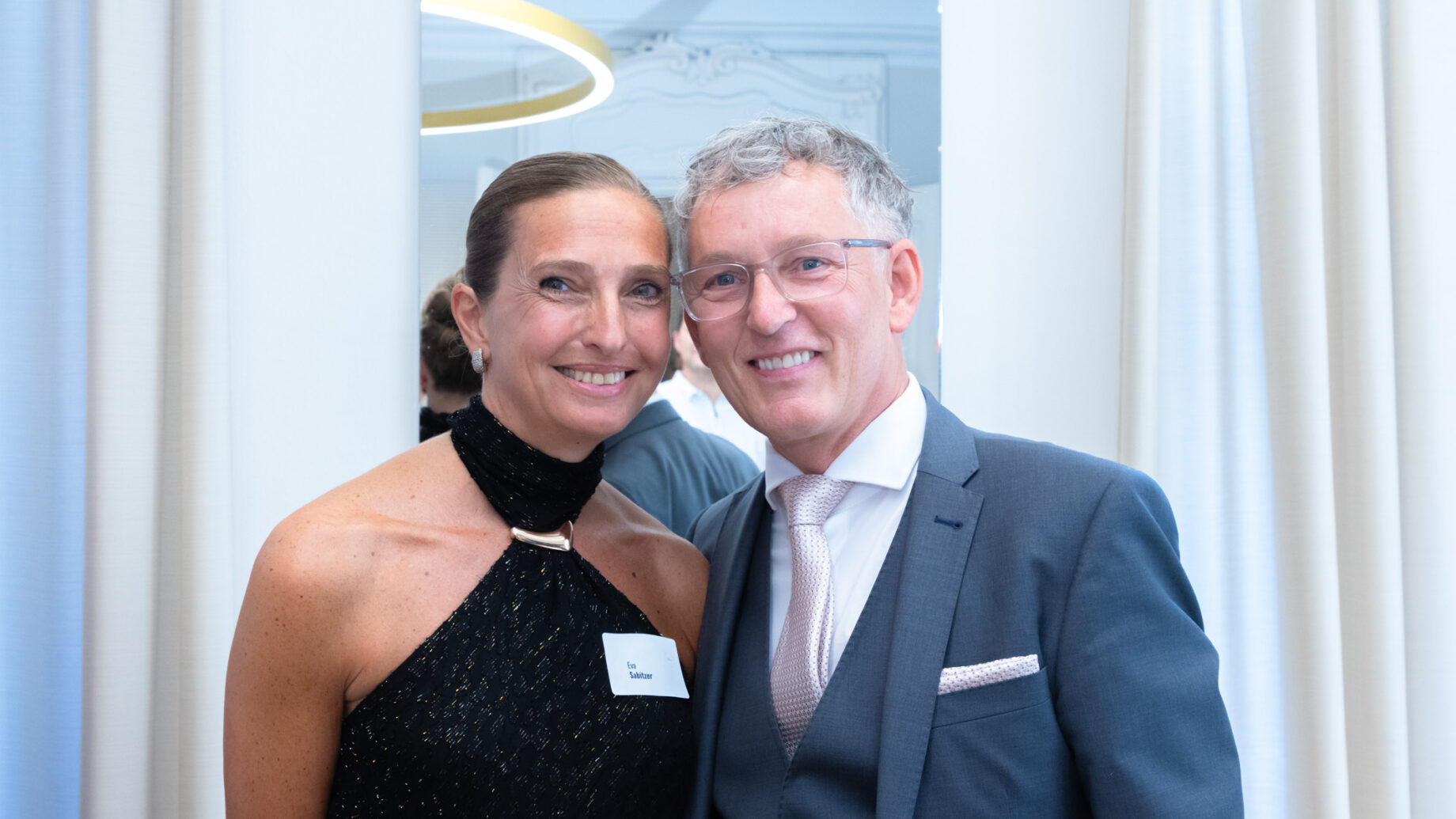 Eine lächelnde Frau in einem schwarzen Kleid und ein Mann im Anzug posieren bei einer Veranstaltung in einer Halle dicht beieinander vor einem weißen Vorhang.