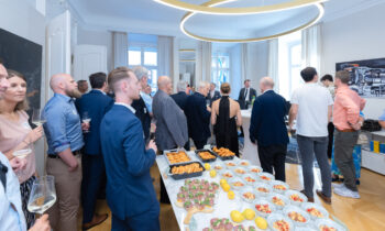 Eine Gruppe formell gekleideter Personen versammelt sich um Tische mit verschiedenen Vorspeisen bei einer hellen Veranstaltung in einem Gebäude. Einige halten Getränke in der Hand, plaudern und mischen sich.
