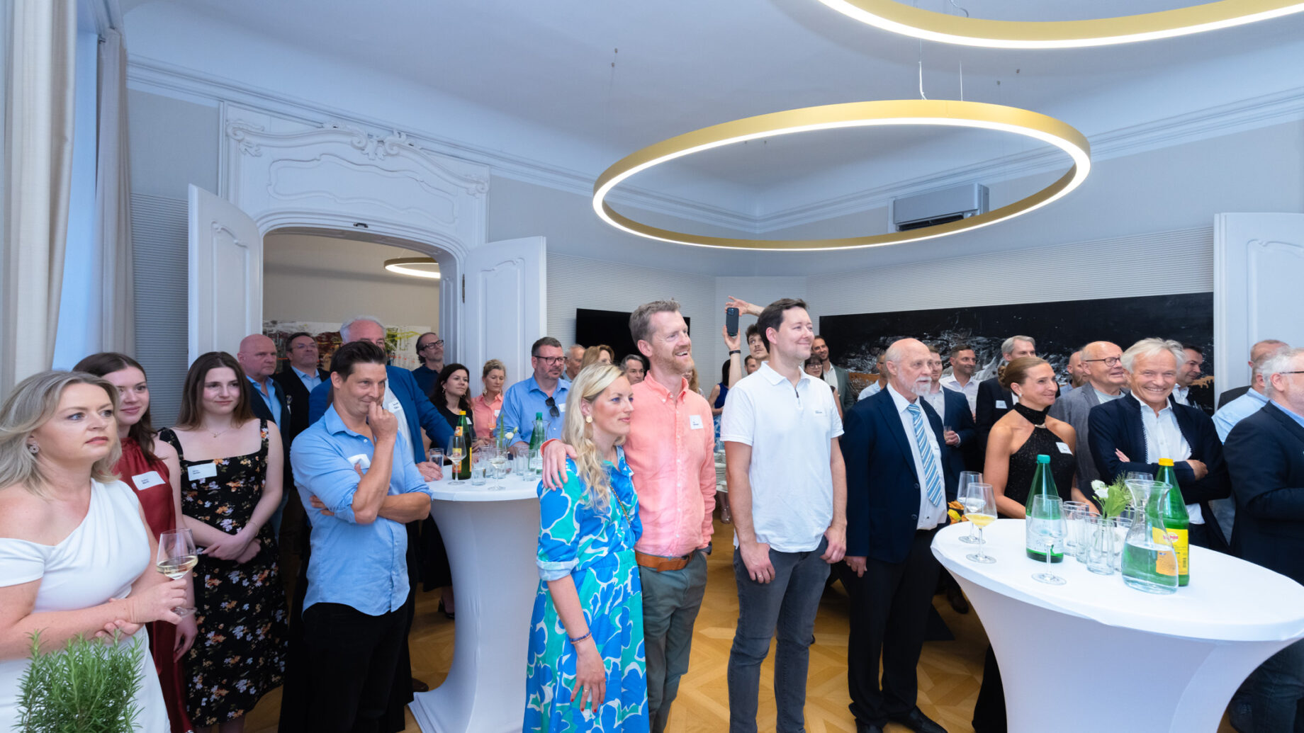 Eine Gruppe von Menschen steht und sitzt bei einer formellen Veranstaltung um Cocktailtische herum und hört in einem hellen Raum mit moderner Beleuchtung aufmerksam zu.