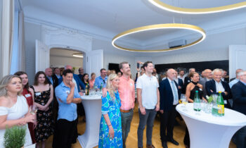 Eine Gruppe von Menschen steht und sitzt bei einer formellen Veranstaltung um Cocktailtische herum und hört in einem hellen Raum mit moderner Beleuchtung aufmerksam zu.