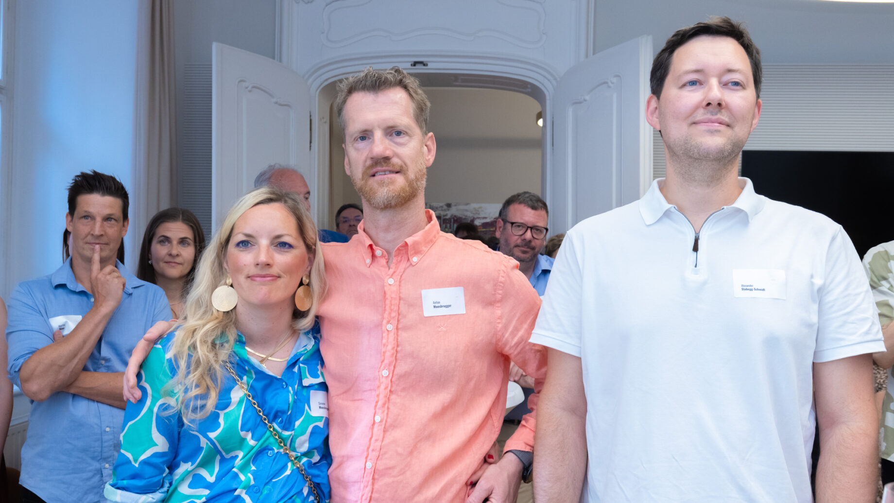 Eine Gruppe von Personen, einige mit Namensschildern, steht in einem Innenraum bei einer Veranstaltung. Drei Personen im Vordergrund posieren gemeinsam, während andere von hinten zusehen.