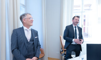 Zwei Männer in Anzügen stehen in einem hellen Raum mit großen Fenstern, einer lächelt, der andere spricht; auf dem Tisch stehen Weingläser und ein Laptop.