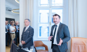 Zwei Männer in Anzügen stehen vor einem Fenster, einer spricht, während der andere zuhört, während im Hintergrund eine Gruppe von Menschen zusieht.