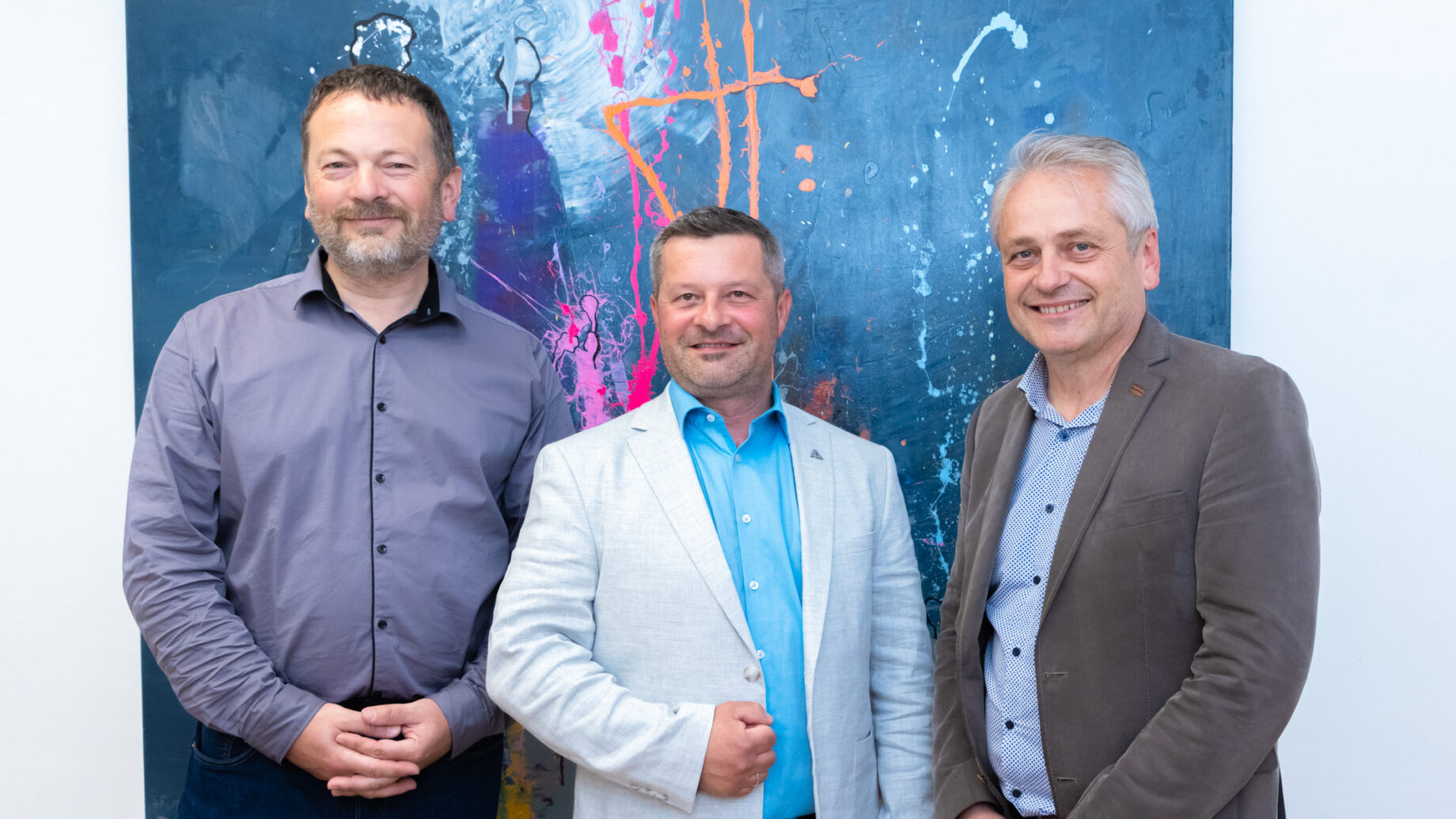 Drei Männer in Geschäftskleidung stehen lächelnd vor einem farbenfrohen abstrakten Gemälde an einer Wand.