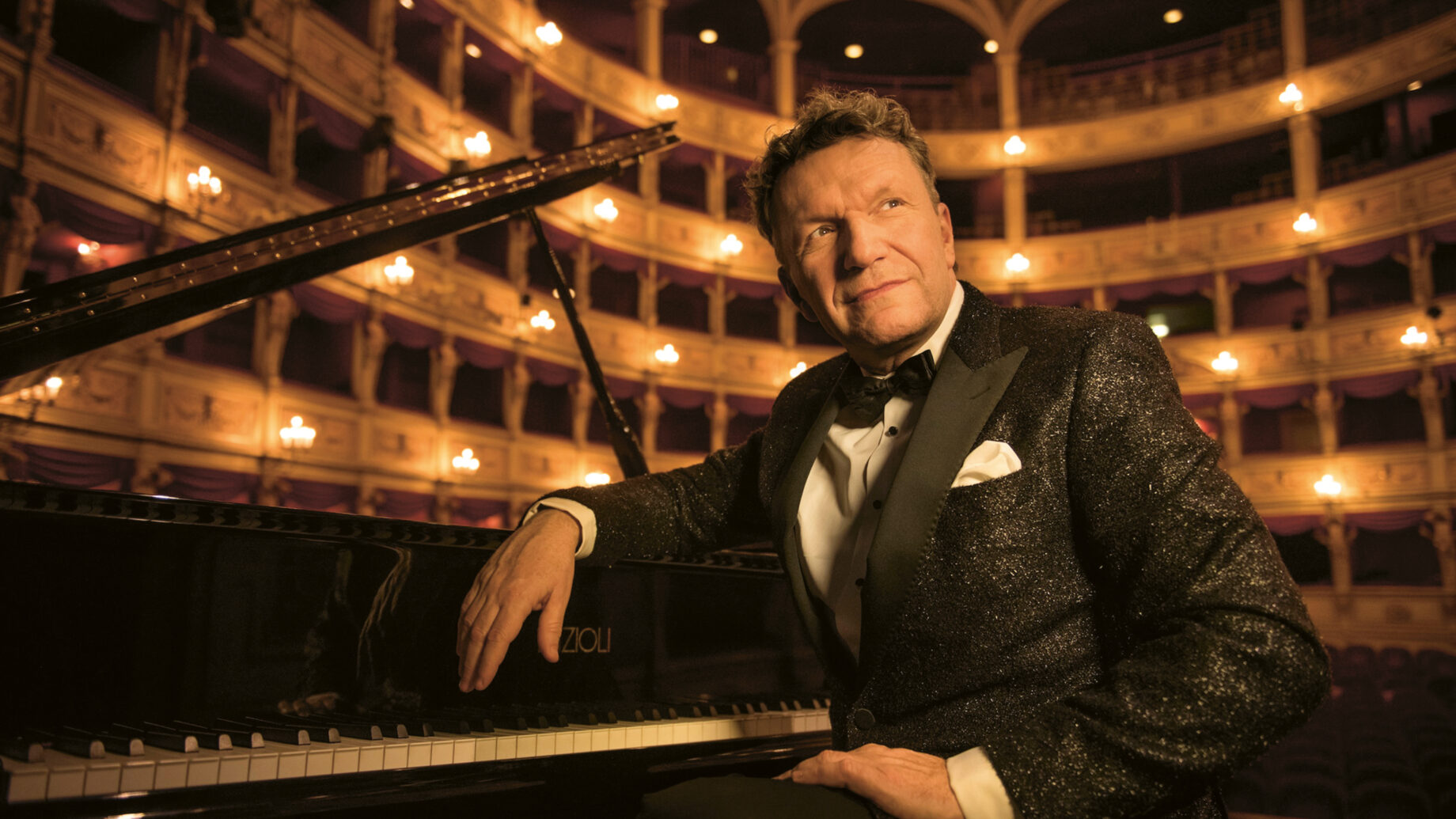 Ein Mann im Smoking sitzt an einem Flügel auf einer Theaterbühne, im Hintergrund sind elegante Balkone und eine warme Beleuchtung zu sehen.