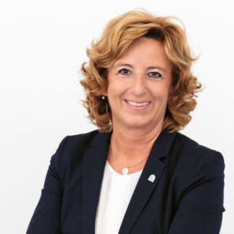 Lächelnde Frau mit lockigem blondem Haar, die einen dunklen Blazer über einem weißen Oberteil trägt und vor einem schlichten weißen Hintergrund steht.