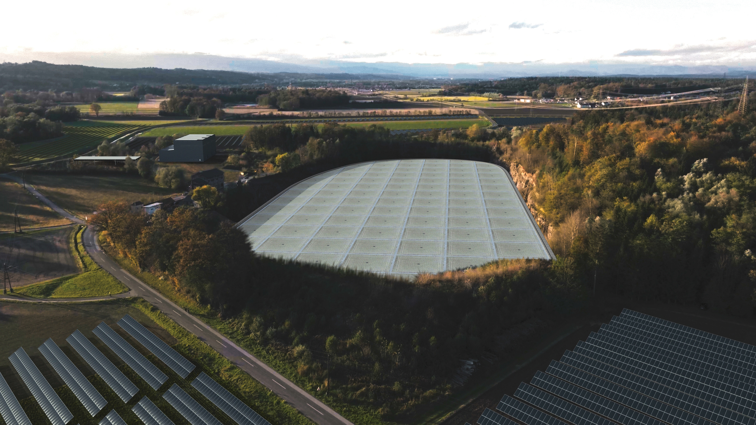 Luftaufnahme eines großen, weißen, rechteckigen Gebäudes, umgeben von Bäumen, Ackerland und Solaranlagen, vor einem teilweise bewölkten Himmel.