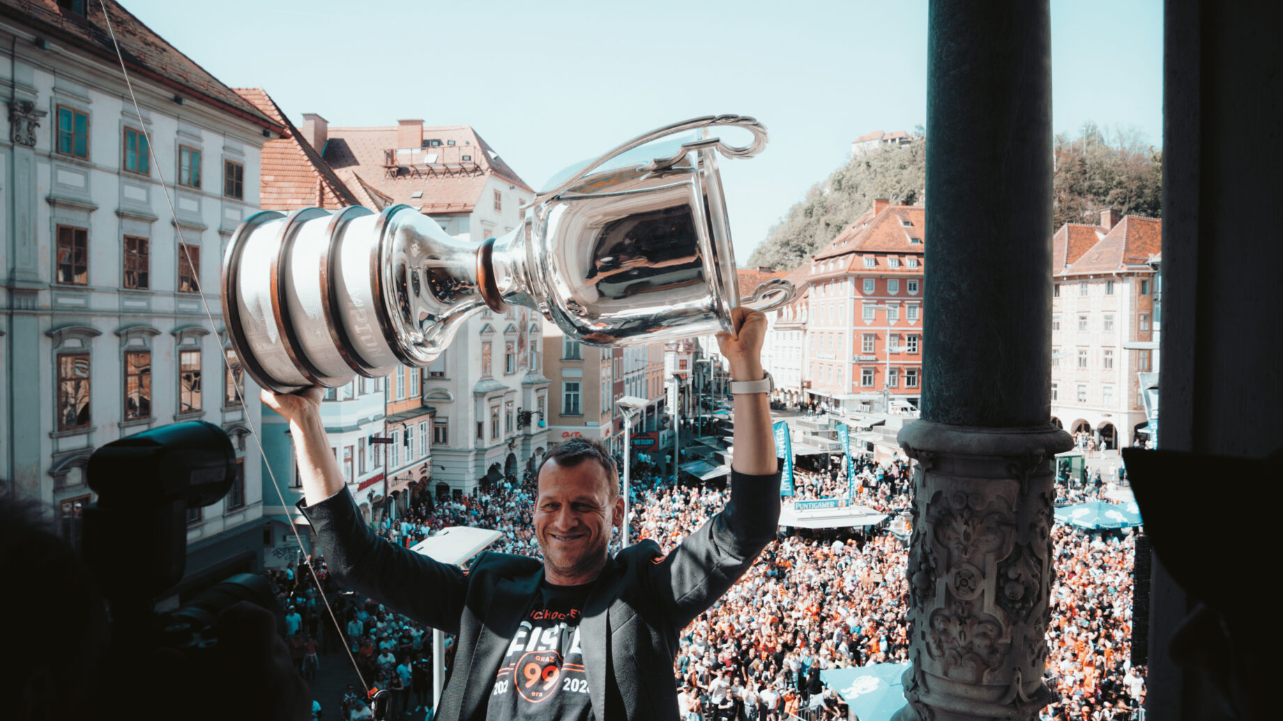 Ein Mann hält auf einem Balkon eine große Trophäe über den Kopf und blickt auf eine jubelnde Menschenmenge auf einem historischen Stadtplatz hinunter.
