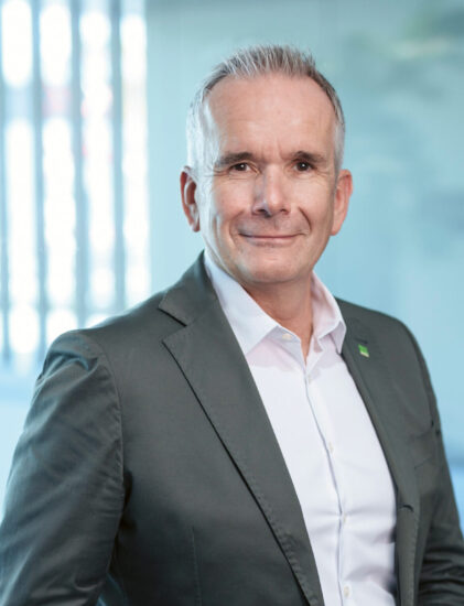 Ein Mann in einem grauen Anzug und einem weißen Hemd steht in einem Raum, lächelt selbstbewusst und hat eine Hand in der Hosentasche. Der Hintergrund ist leicht unscharf.