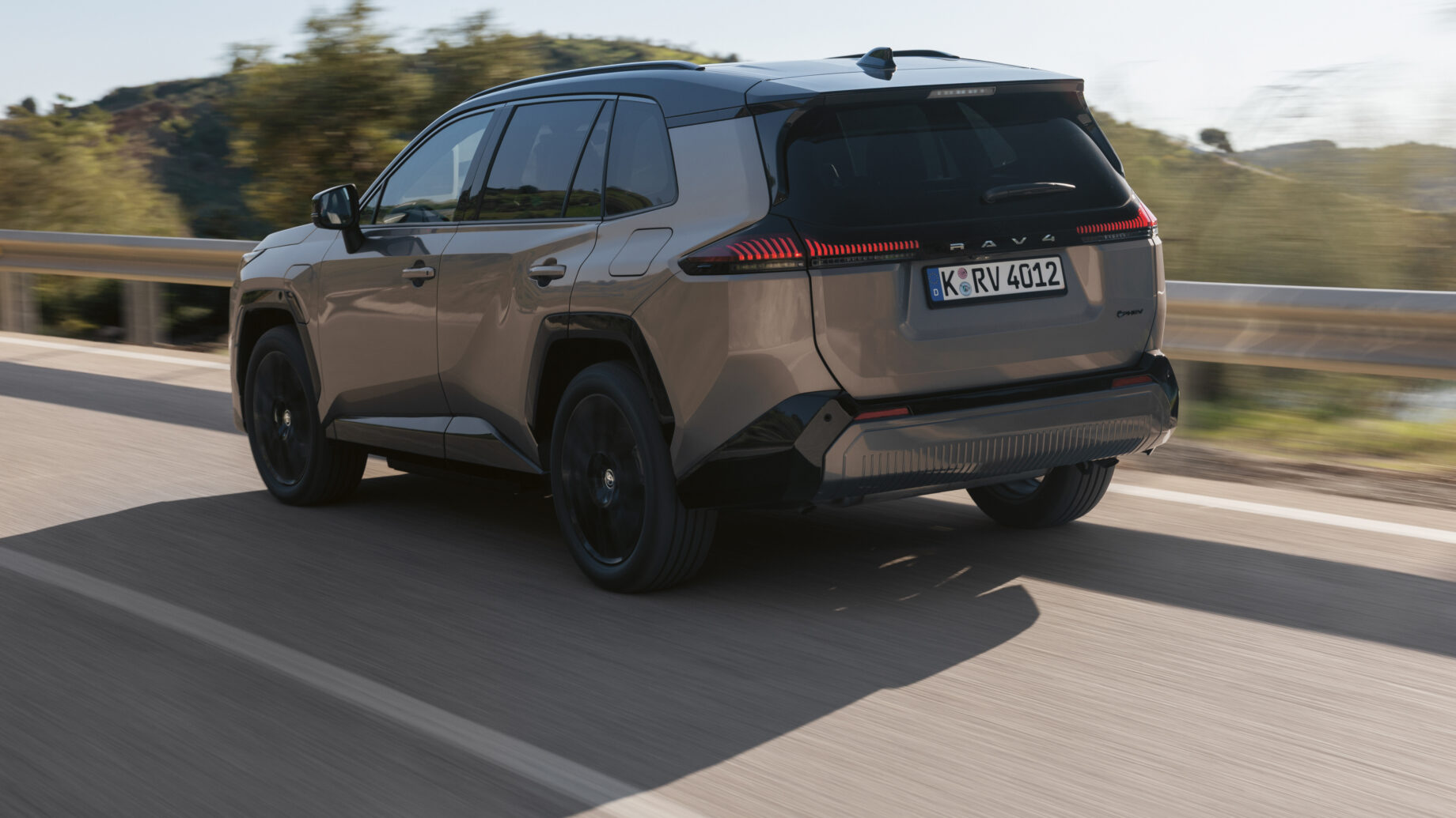 Ein beiger SUV fährt auf einer sonnigen Straße, im Hintergrund sind Bäume und Hügel zu sehen; die Aufnahme stammt von der linken Rückseite.