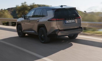 Ein beiger SUV fährt auf einer sonnigen Straße, im Hintergrund sind Bäume und Hügel zu sehen; die Aufnahme stammt von der linken Rückseite.