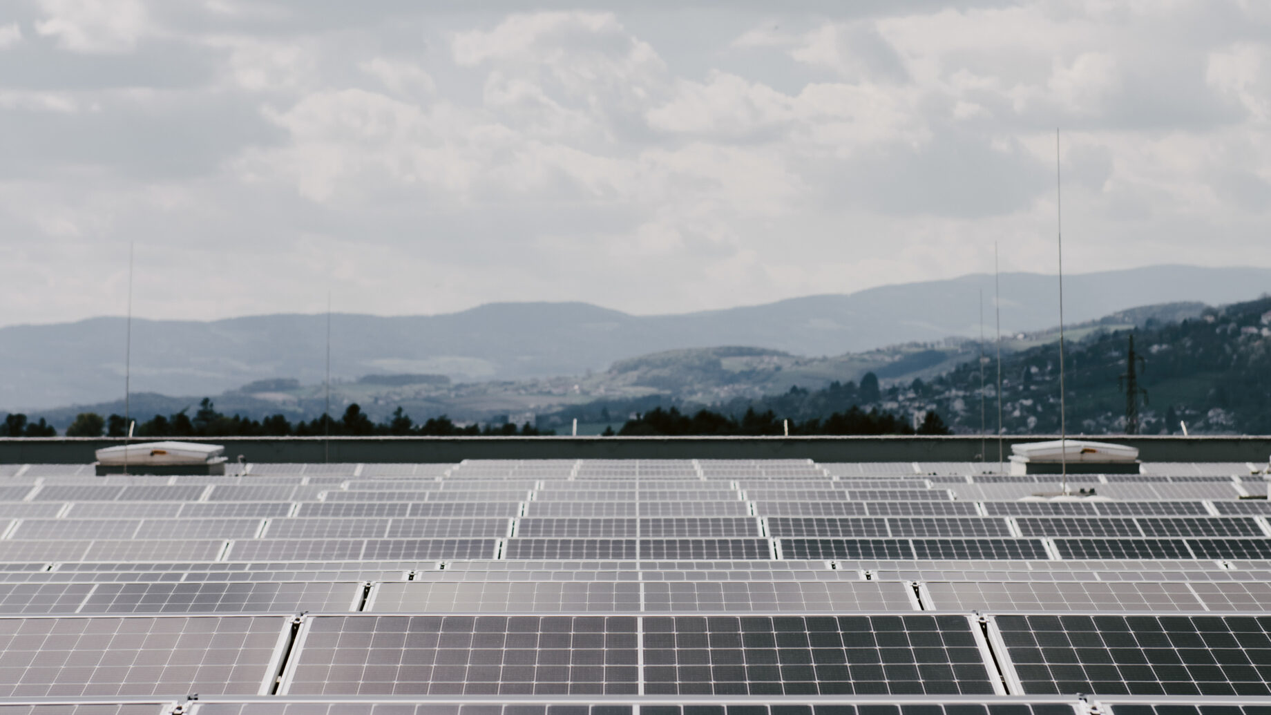 Reihen von Solarmodulen auf einem Dach, im Hintergrund ein bewölkter Himmel und ferne Hügel.