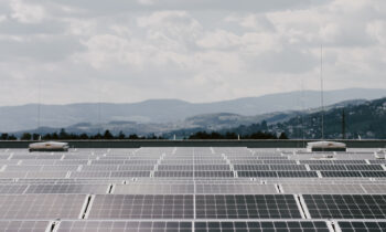 Reihen von Solarmodulen auf einem Dach, im Hintergrund ein bewölkter Himmel und ferne Hügel.