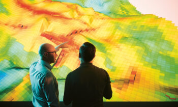 Zwei Personen unterhalten sich über eine farbenfrohe 3D-Topografiekarte, die auf einem großen Bildschirm angezeigt wird, wobei eine Person auf die Karte zeigt.