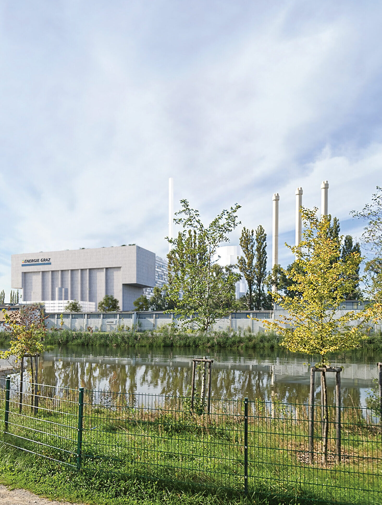 Industriegebäude mit hohen Schornsteinen hinter einem Teich, grünen Bäumen, einer Rasenfläche und einem Zaun im Vordergrund unter einem teilweise bewölkten Himmel.
