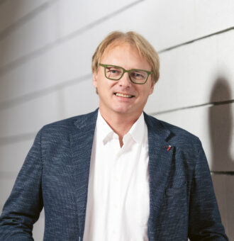 Ein Mann mit Brille und blauem Blazer steht vor einer weißen, strukturierten Wand und lächelt.