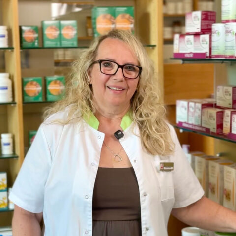 Eine lächelnde Frau in einem weißen Kittel steht in einer Apotheke, umgeben von Regalen mit Gesundheitsprodukten und Nahrungsergänzungsmitteln.