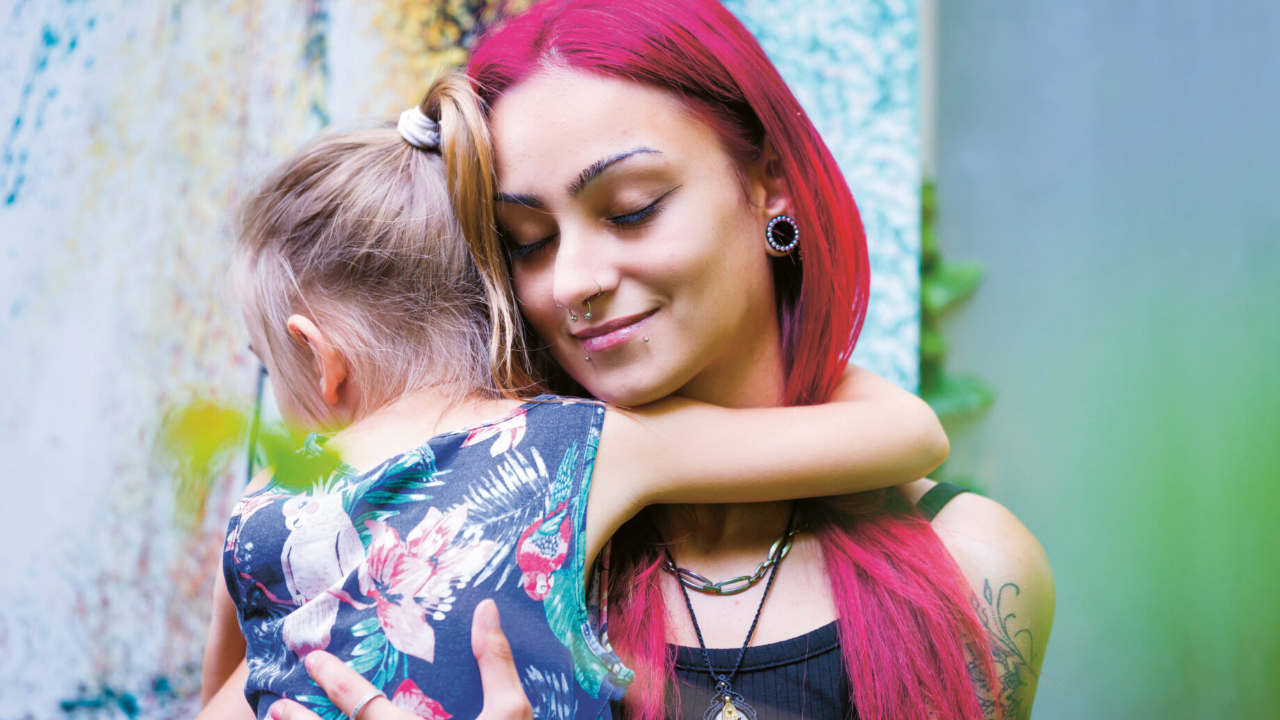 Eine Frau mit langen rosa Haaren umarmt ein junges Mädchen mit Pferdeschwanz. Beide wirken ruhig und zufrieden, während sie draußen vor einer bunten Wand stehen.