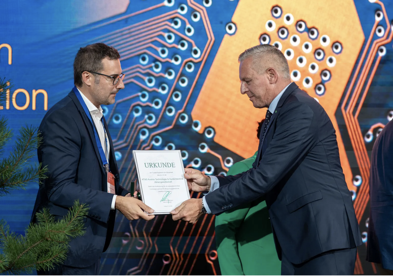 Zwei Männer in Anzügen überreichen sich auf der Bühne eine Urkunde, im Hintergrund ist das Leiterplatten-Design des steirischen WKO-Wirtschaftsbarometers zu sehen.